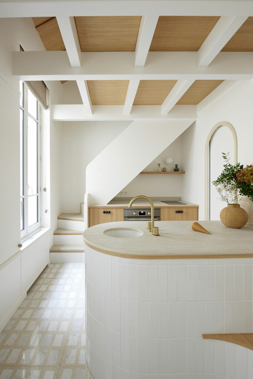 Cocina moderna con azulejos blancos y acentos de madera, con una isla curva con un fregadero, plantas en el mostrador y un horno debajo de las escaleras. Las escaleras conducen, y una gran ventana deja en la luz natural.