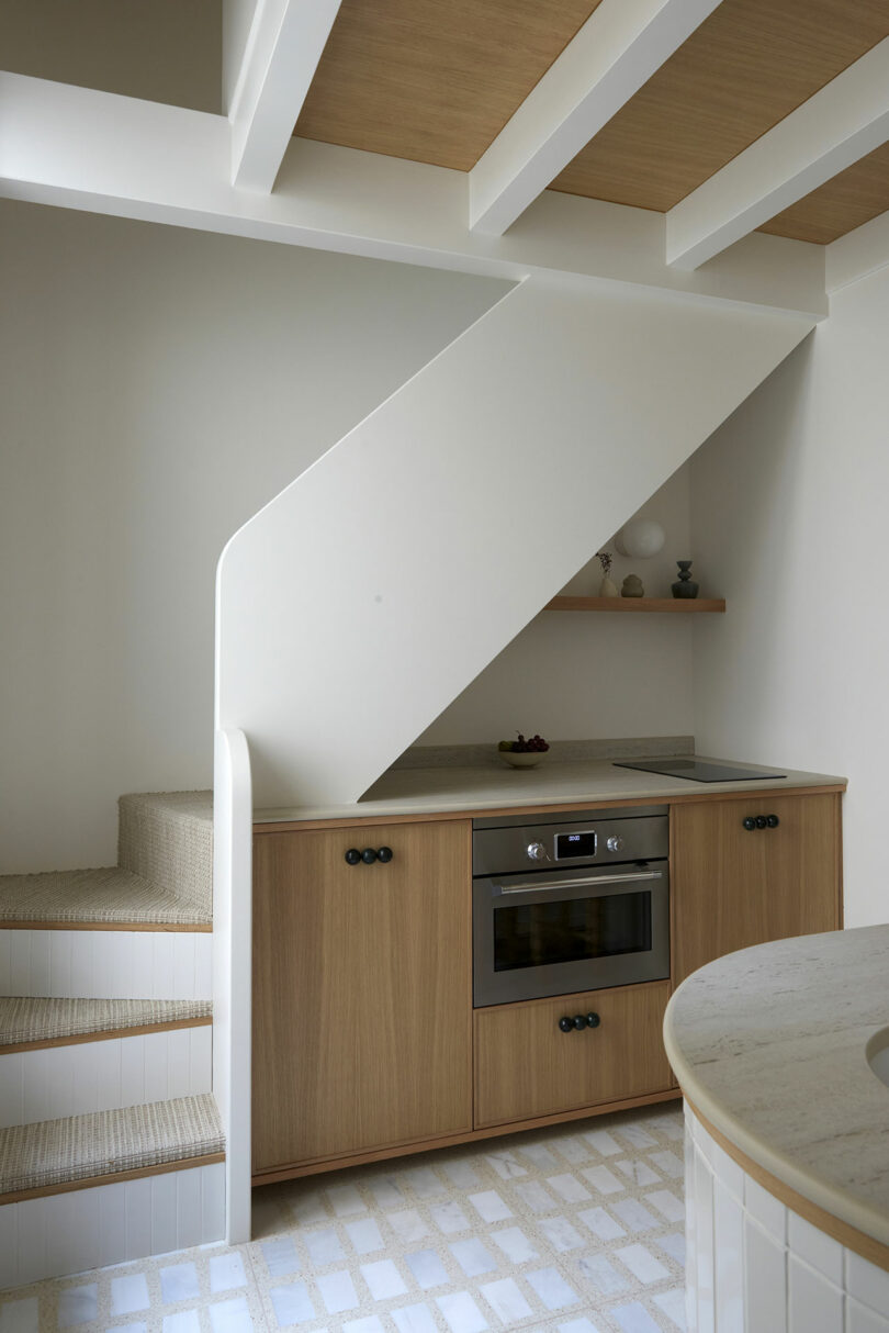 Cocina moderna con gabinetes de madera debajo de una escalera, con un horno incorporado, una decoración minimalista en un estante y pisos de azulejos.