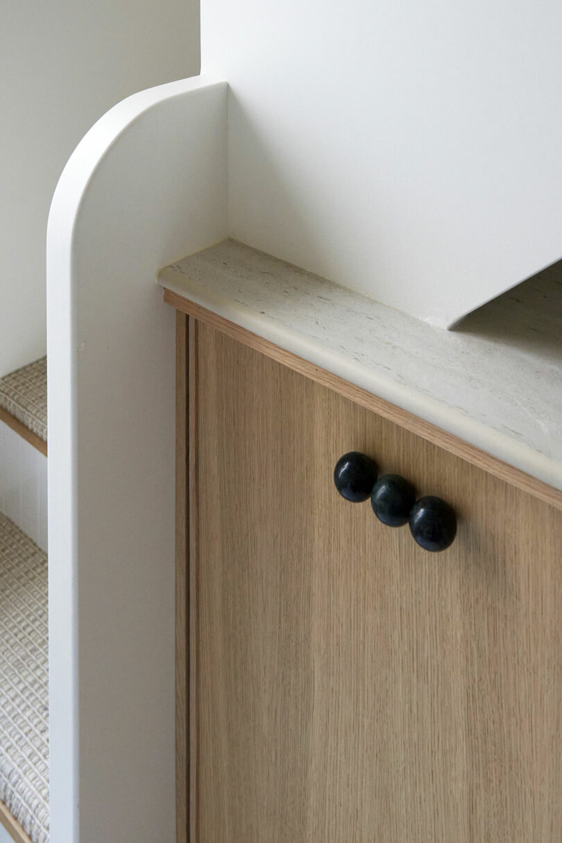 Close-up of a wooden cabinet with three round black handles, topped with a light stone surface and adjacent to a staircase with carpeted steps.