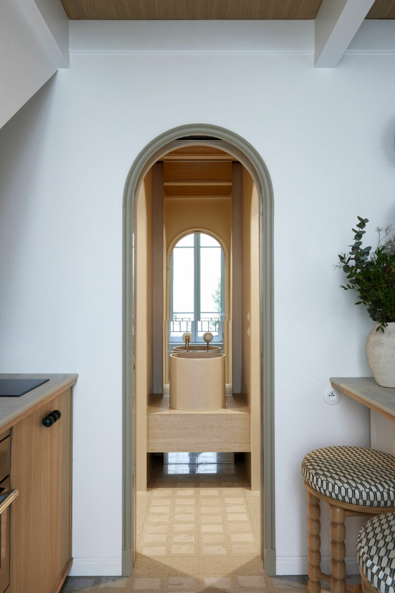 Puerta arqueada que conduce a una cocina minimalista con gabinetes de madera, una isla central con taburetes, un jarrón con vegetación y una ventana en el fondo.