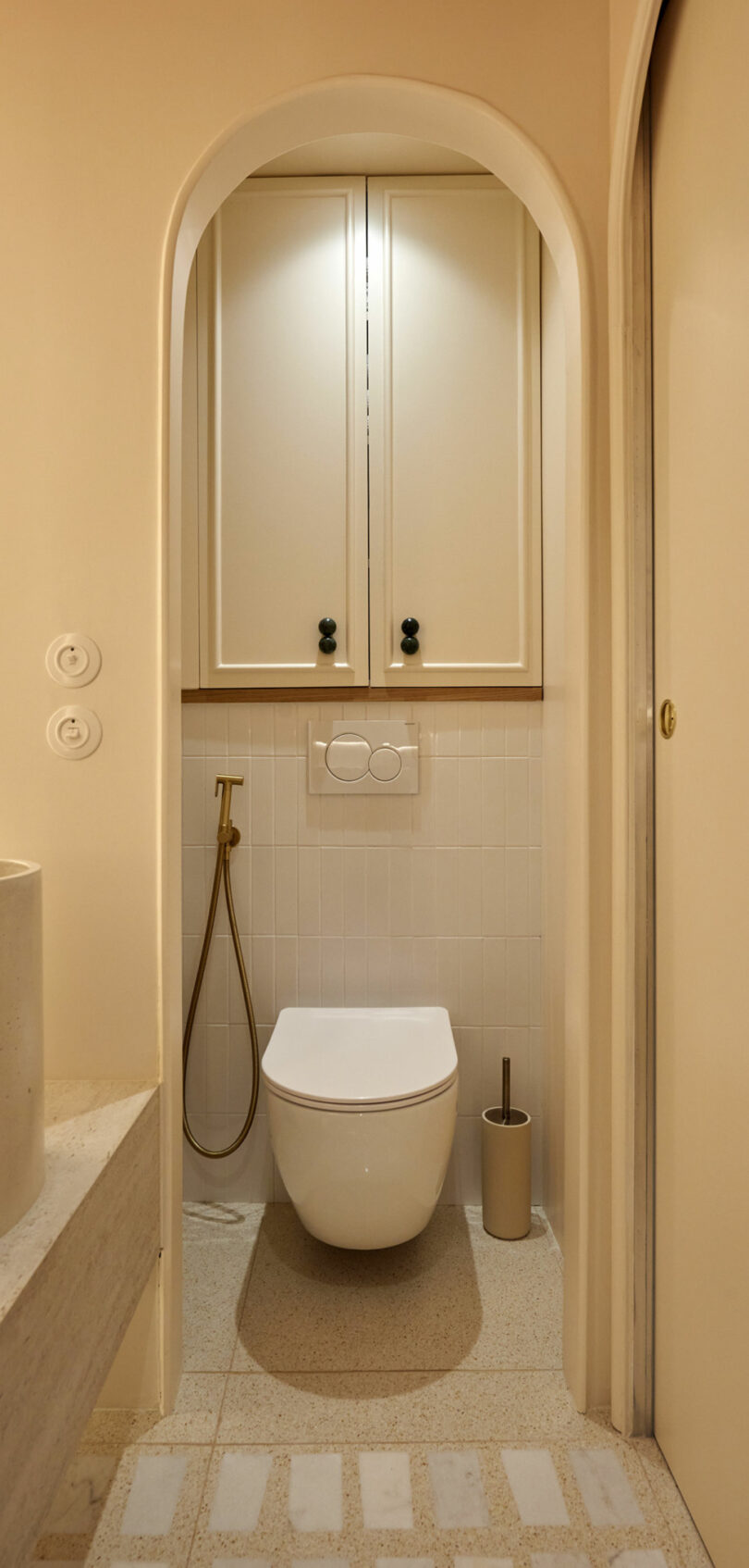 Minimalist bathroom with a modern toilet, wall-mounted flush, a small cabinet above, and a bidet spray beside it. The neutral-toned walls and floors create a warm atmosphere.