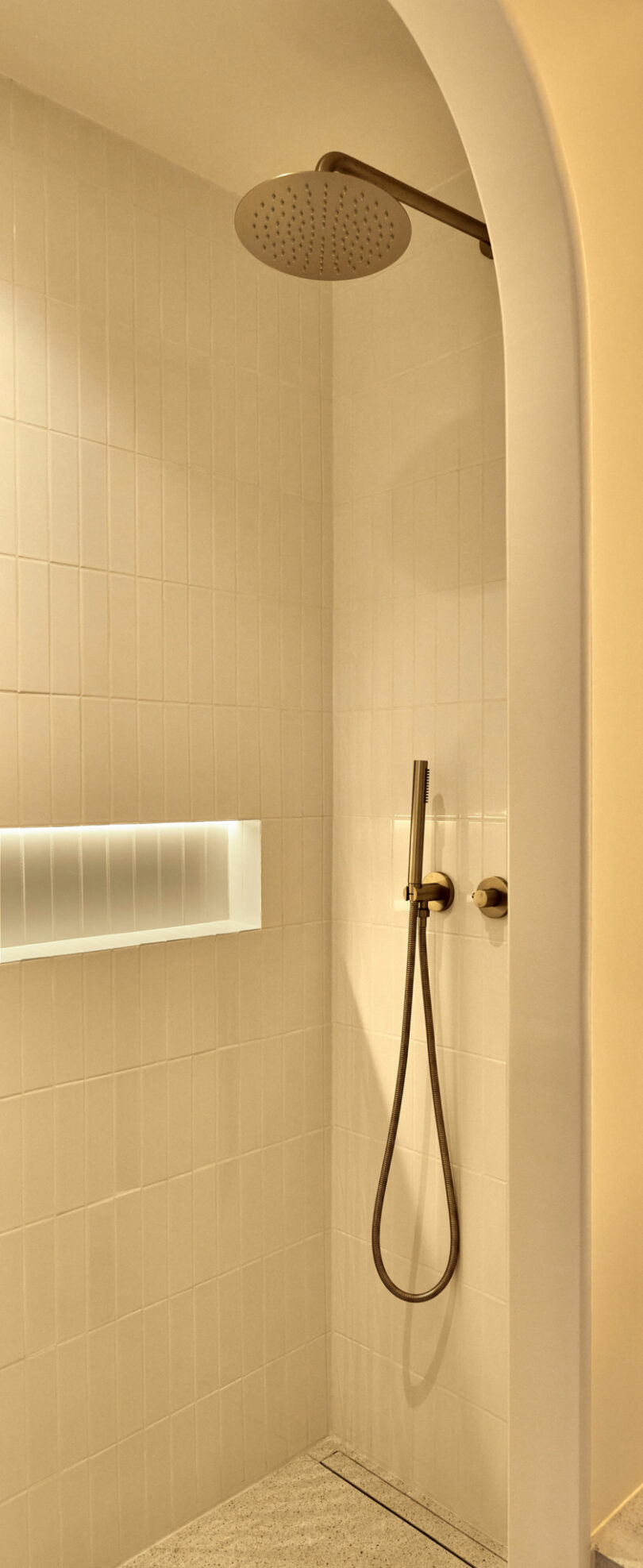 Minimalist shower with a large overhead rain showerhead, a handheld shower wand, and white tiled wall. Built-in illuminated shelf holds toiletries.