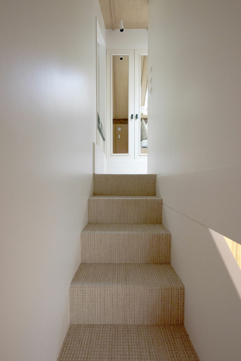 Una escalera estrecha y alfombrada conduce a un rellano con un espejo de pared y un techo de madera en un interior moderno y minimalista.