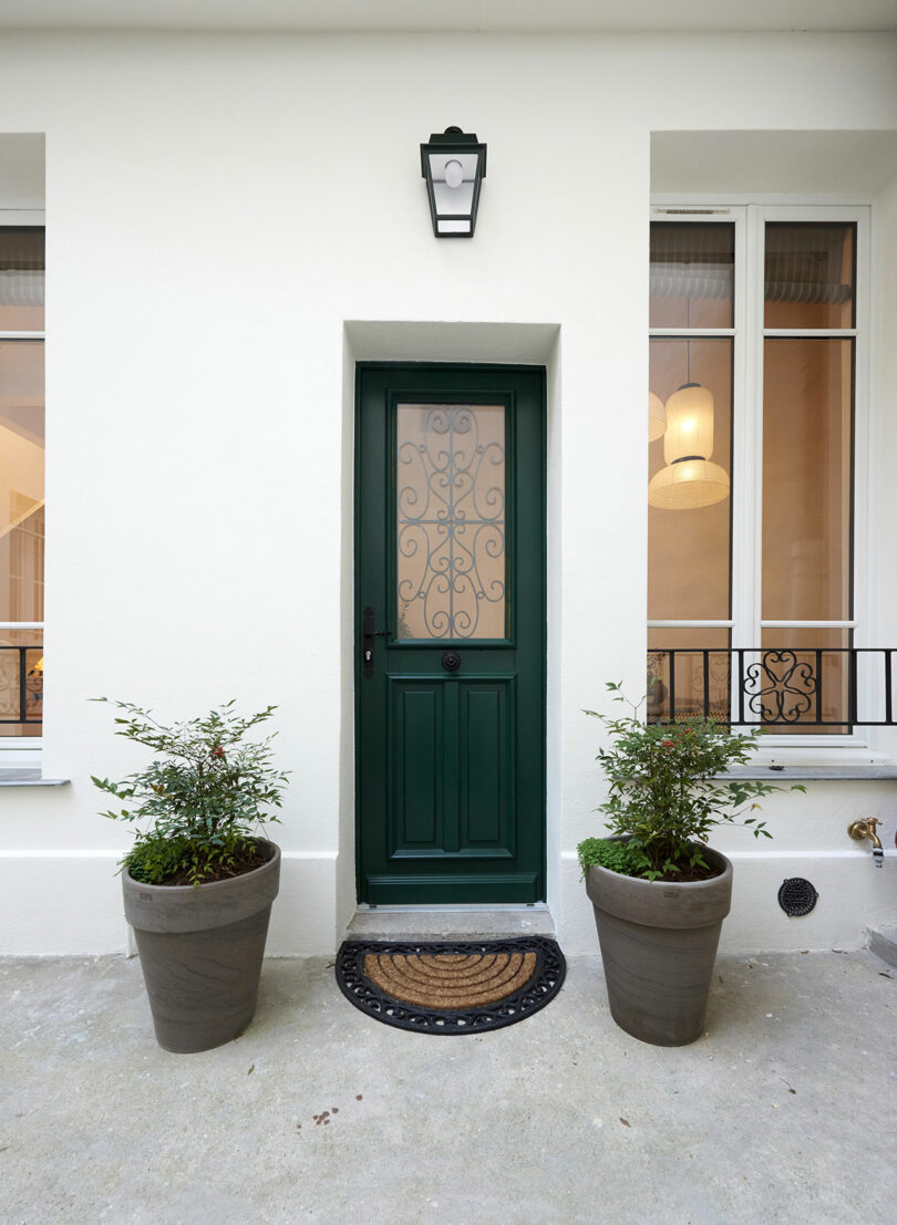 Puerta verde con panel de vidrio adornado flanqueado por dos plantas en macetas en una fachada de edificios de color claro, debajo de una lámpara de exterior negra.