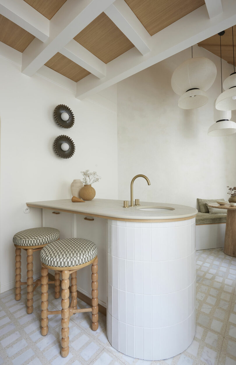 Modern kitchen with a curved, tiled island, wooden countertop, and two patterned stools. Ceiling has wooden beams; spherical light fixtures hang above. Walls have artistic decor pieces.