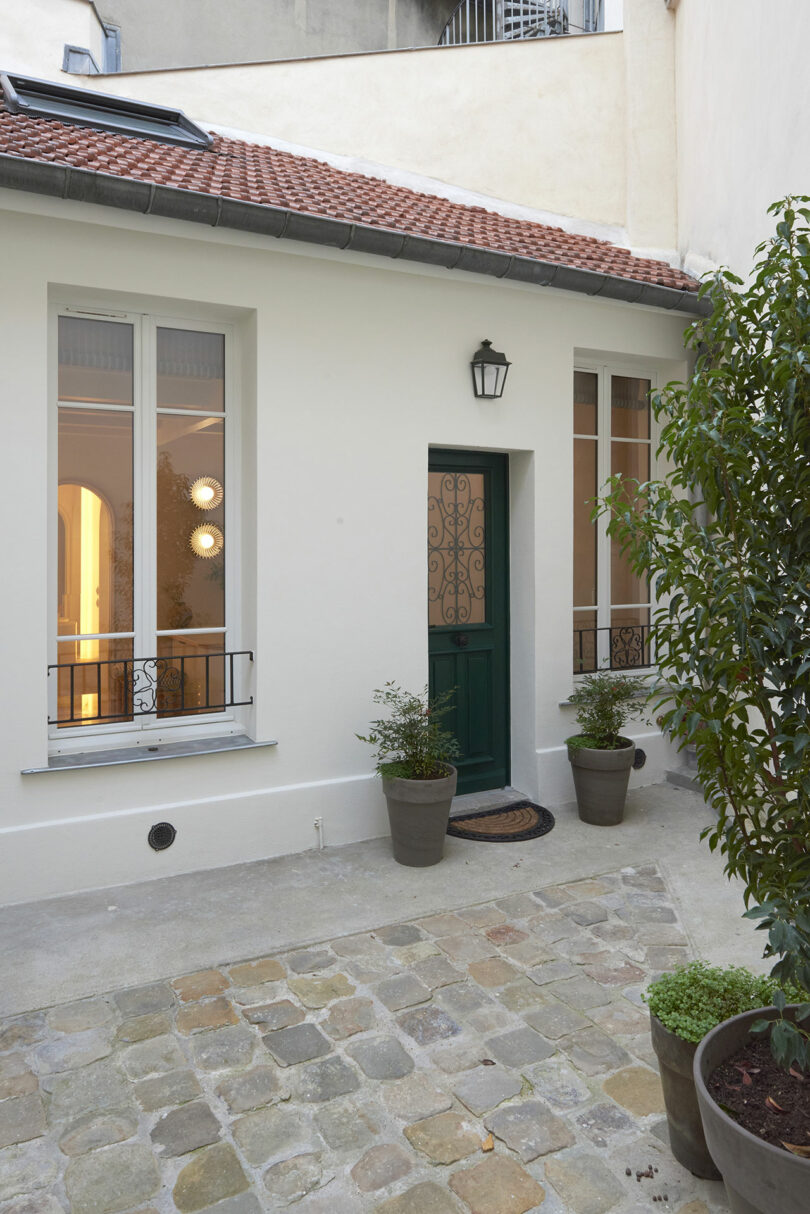 Patio pequeño con pisos de adoquines, plantas en macetas y una fachada de edificio blanco. Puerta verde con diseño de hierro forjado y dos ventanas. Linterna de pared sobre la puerta.