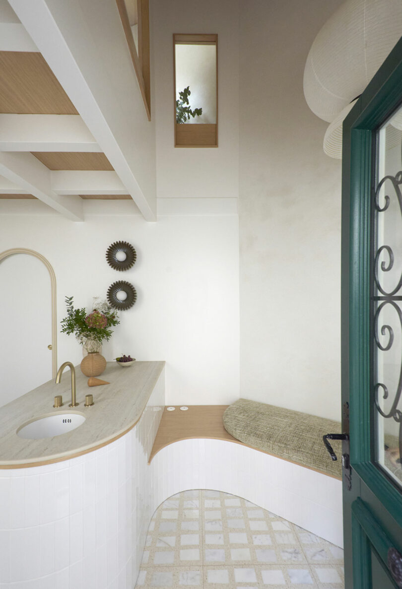 A minimalist bathroom featuring a white curved countertop with a sink, decorative items, a cushioned bench, and circular wall mirrors. A green door with glass panels is partially visible.