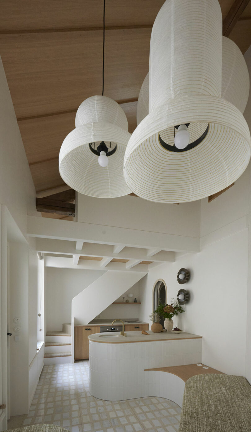 Cocina minimalista con isla curva blanca, techo de madera y grandes luces colgantes blancas. Una escalera conduce arriba, y una pequeña planta se encuentra en una encimera.