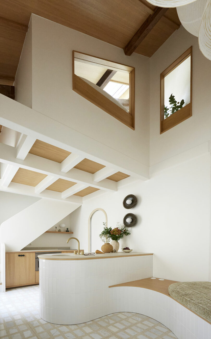 Una cocina moderna y minimalista con un techo alto, paredes blancas, acentos de madera y ventanas geométricas. Cuenta con un mostrador curvo, asientos incorporados y un piso ligero de baldosas.