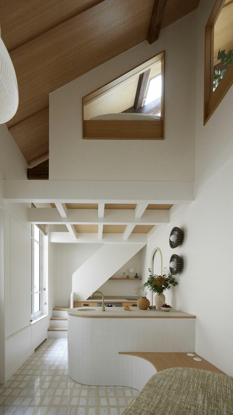 Modern kitchen with white walls, wood accents, and high slanted ceiling. Features include a countertop with vase, built-in oven, and large window. Decorative items hang on the wall.