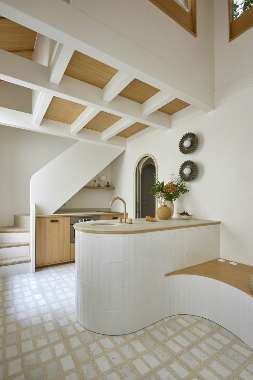 Interior de cocina moderno con isla de azulejos blancos curvos, acentos de madera y decoración contemporánea. Cuenta con una escalera, una puerta arqueada y un diseño de techo geométrico.