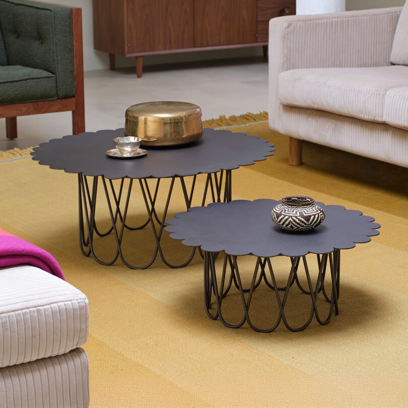 Two scalloped-edge black coffee tables with metal bases on a yellow rug, adorned with decorative items, are in a living room with a green chair and beige sofas.