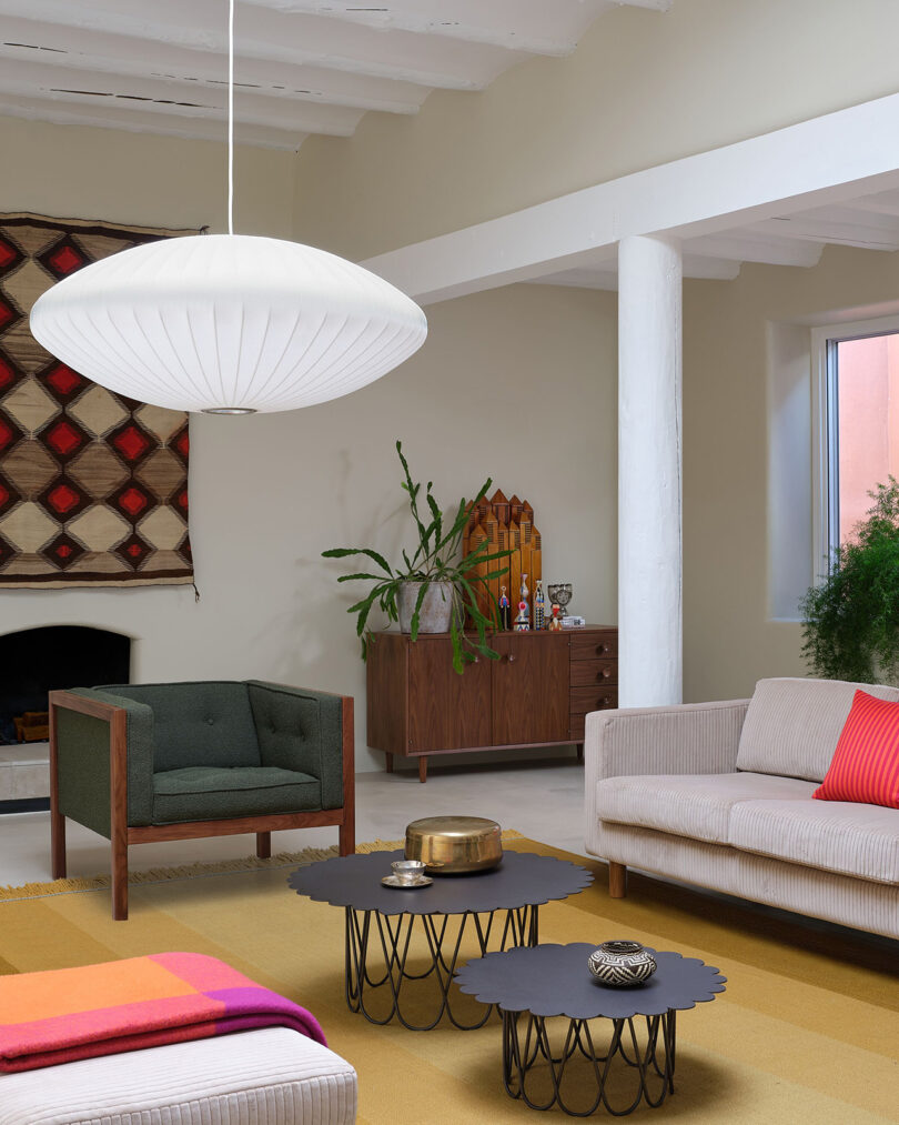 Modern living room with a white pendant light, green armchair, beige sofa with red cushion, geometric wall art, and nested black tables on a yellow rug. Cabinet and plants in the background.