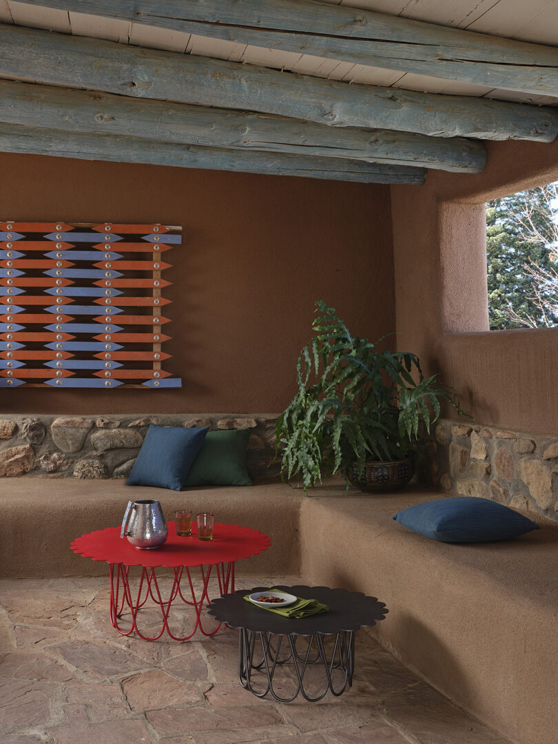 Covered patio with two round wireframe tables, one red and one black, a built-in bench with cushions, and a plant. Geometric wall art and a window view of snowy trees in the background.