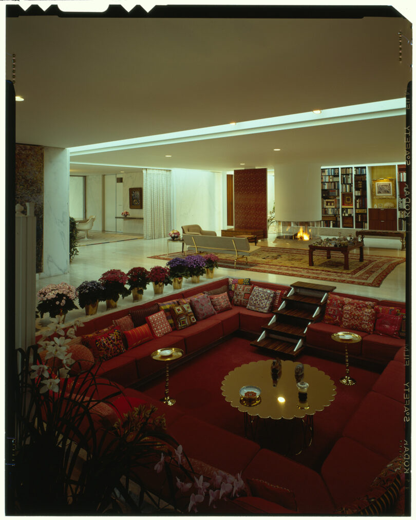 Large, elegant living room with sunken seating area, red couches, gold tables, and abundant floral arrangements. Background features a fireplace, bookshelves, and a lounge chair.