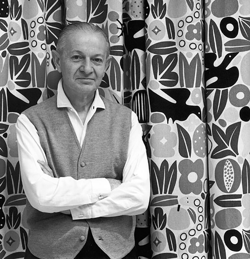 An individual stands with arms crossed in front of a patterned curtain with abstract shapes and flowers.