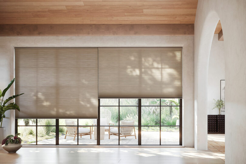 A room with large windows features closed blinds, cream walls, and wooden flooring. Outdoors, there are two wicker chairs on a patio with greenery in the background.