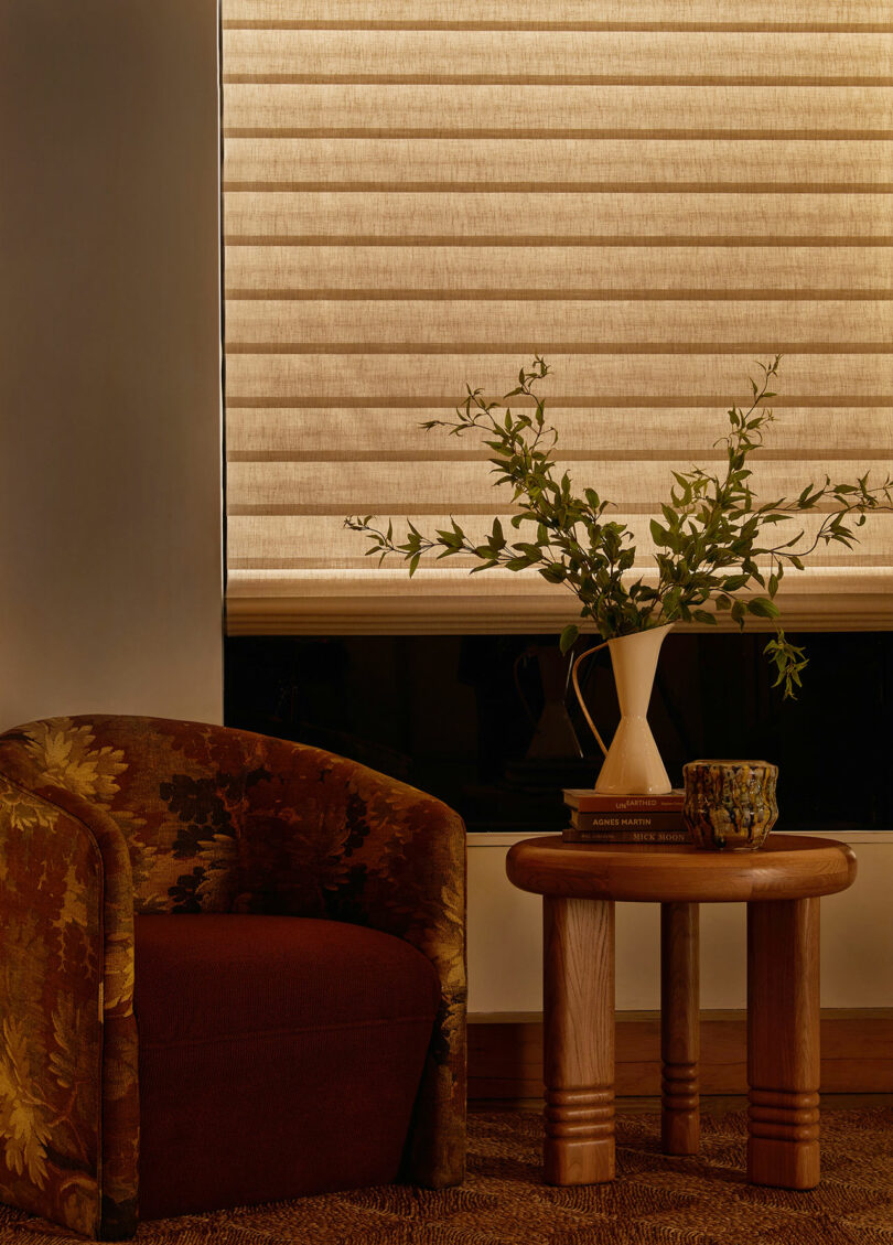 A cozy room corner with a floral-patterned armchair, a small wooden table with a vase of greenery, and a covered window with beige blinds.