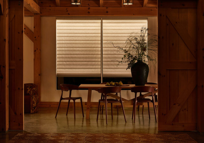 A dimly lit dining room features a wooden table with four chairs. A large plant in a black vase sits on the table. Beige curtains cover the windows, and the walls have wooden beams.