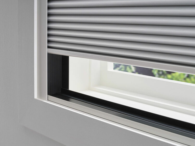 White window with a partially open roller blind, revealing a view of greenery outside. The blind is silver with a horizontal slat design.