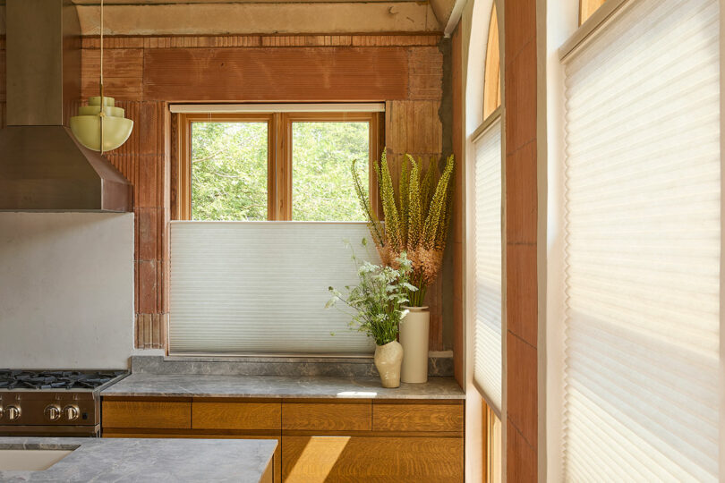 Modern kitchen with wooden cabinets and marble countertop. Large window with white blinds, green vase with flowers, and stainless steel range hood. Natural light illuminates the space.