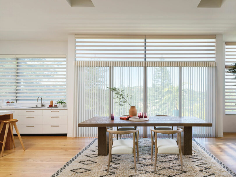 Modern dining room with a wooden table, chairs, and a light rug. Large windows with horizontal blinds allow natural light. Kitchen area with sink and stool visible on the left.