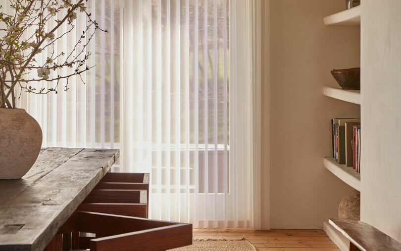 Minimalist dining area with a wooden table and chairs, large potted plant, sheer curtains, and a wall shelf with books and decor. Natural light filters in through the window.