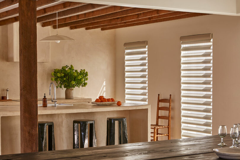 Modern kitchen with an island featuring black stools, a vase of green flowers, and a bowl of oranges. Three striped windows allow sunlight in, and a wooden chair is against the wall.