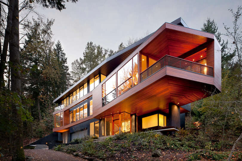 Modern Angular House con grandes ventanas y acentos de madera, ubicado en un área boscosa. La casa está elevada en pilares y rodeada de árboles y vegetación.