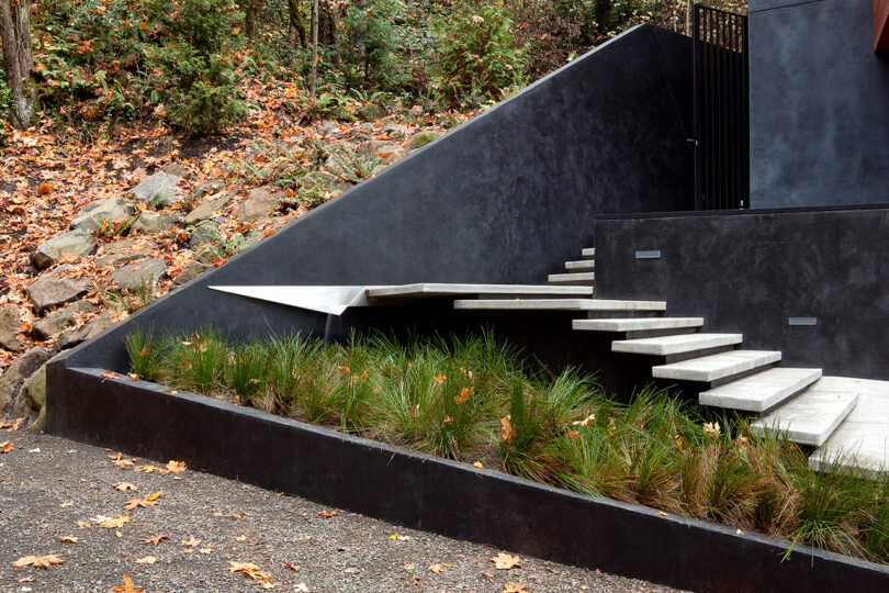 Los pasos de concreto con un diseño minimalista conducen a una pendiente ajardinada con hierba y hojas caídas, junto a una pared de retención oscura.