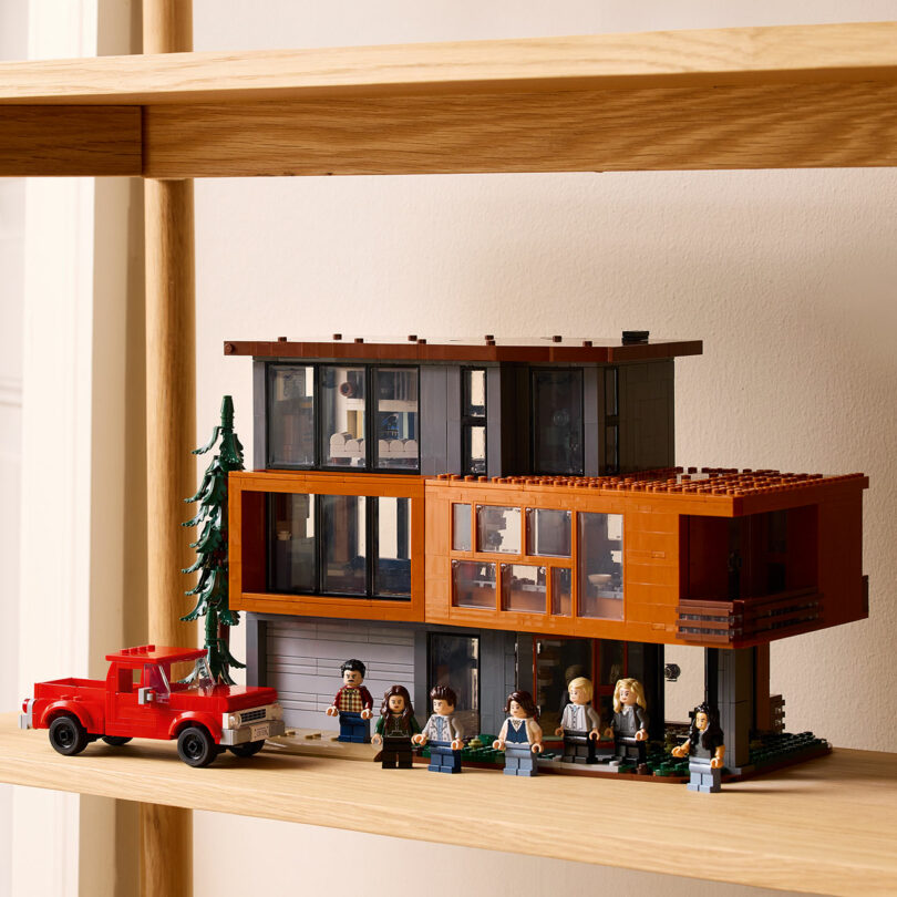 Se muestra un modelo LEGO detallado de una moderna casa de dos pisos con grandes ventanas en un estante de madera. En el frente, hay una camioneta roja de Lego y varias minifiguras.