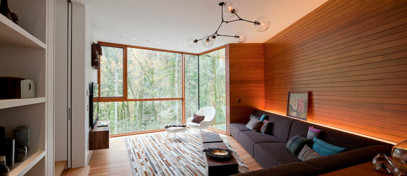 Modern living room with large windows, wooden wall panels, a dark sectional sofa, decorative pillows, a white lounge chair, and a unique ceiling light fixture.