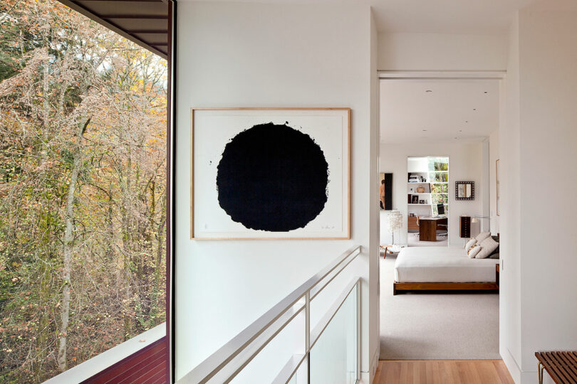 Hallway in a modern home with a large black circle artwork on the wall. The room ahead has a bed and large windows overlooking trees.