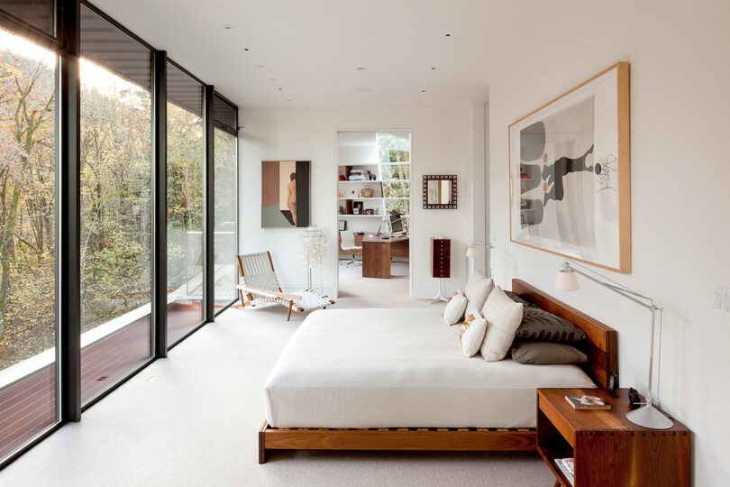 Dormitorio moderno con grandes vidrio, una cama de madera y una vista de árboles. Un área abierta adyacente cuenta con una silla y arte de pared.