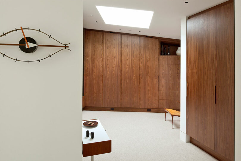 Habitación moderna con gabinetes de madera, un reloj montado en la pared con forma de ojo, una mesa y una claraboya. Paleta de colores neutros con tonos marrones y blancos.