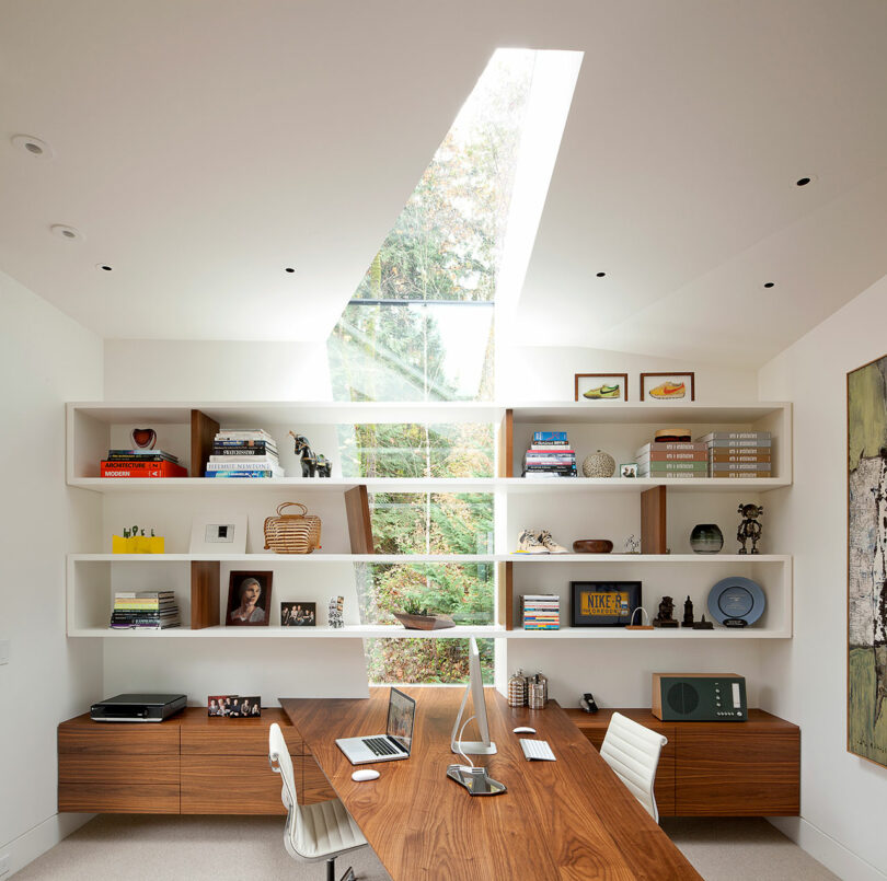 Moderna oficina en casa con un escritorio de madera, computadora y silla. Los estantes montados en la pared muestran libros y artículos decorativos. Una claraboya arriba ilumina el espacio, y una ventana muestra vegetación afuera.