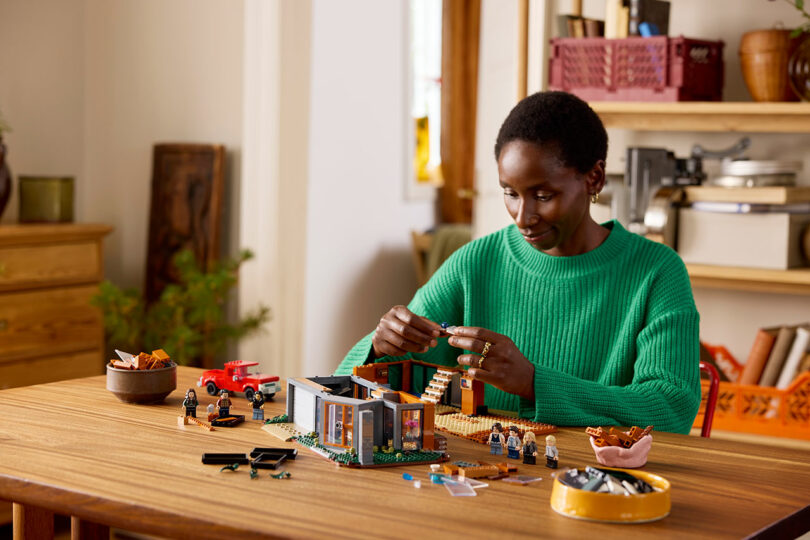 Persona en un suéter verde que ensambla un LEGO en una mesa de madera, rodeada de figuras y piezas en miniatura, con un fondo de estantes y plantas.