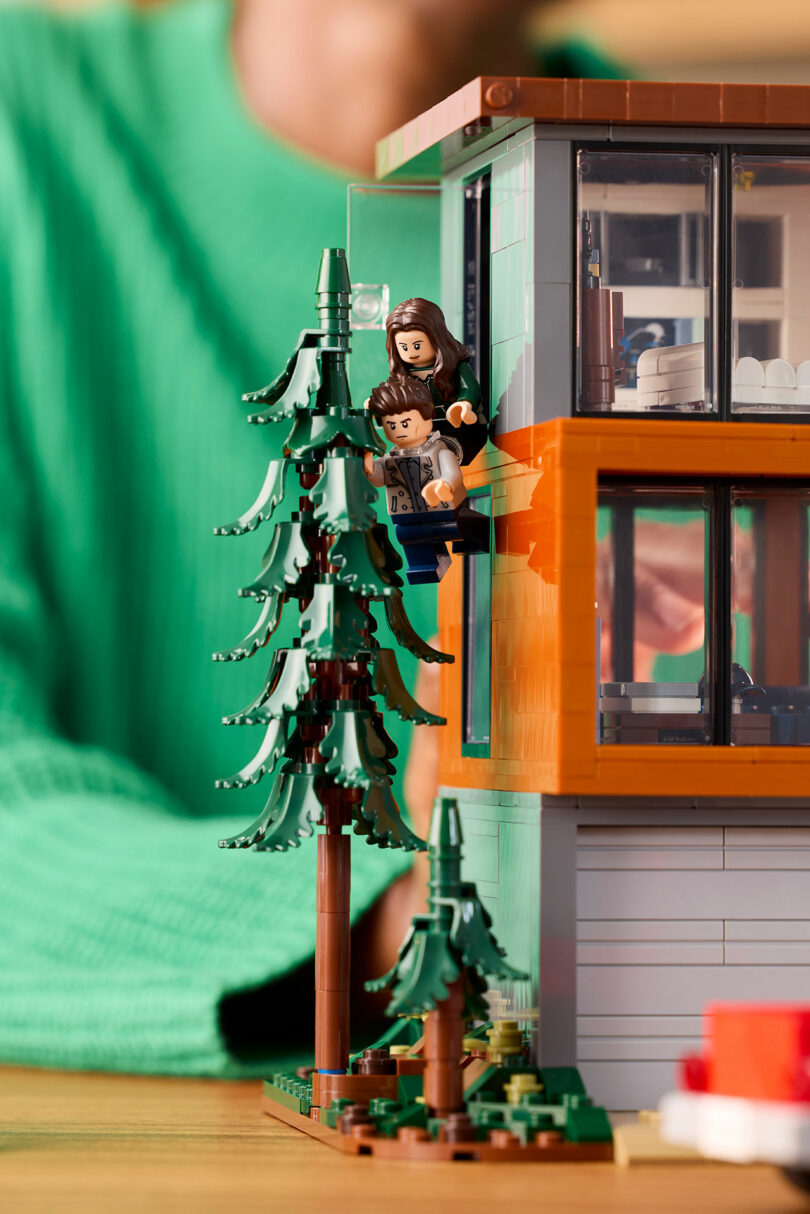 Lego minifigures cling to a window ledge of a large, modern house, surrounded by pine trees.