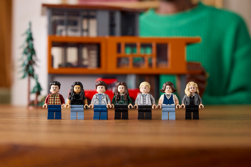 Seven Lego minifigures are lined up on a table in front of a detailed building model.