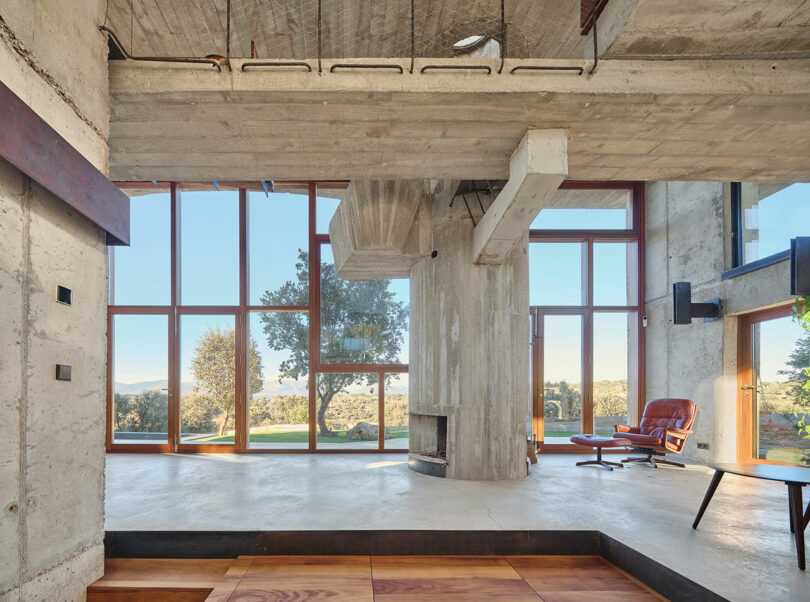 Spacious modern interior with concrete walls, large windows, and a view of trees outside. Features a fireplace, wooden flooring, and a red lounge chair.