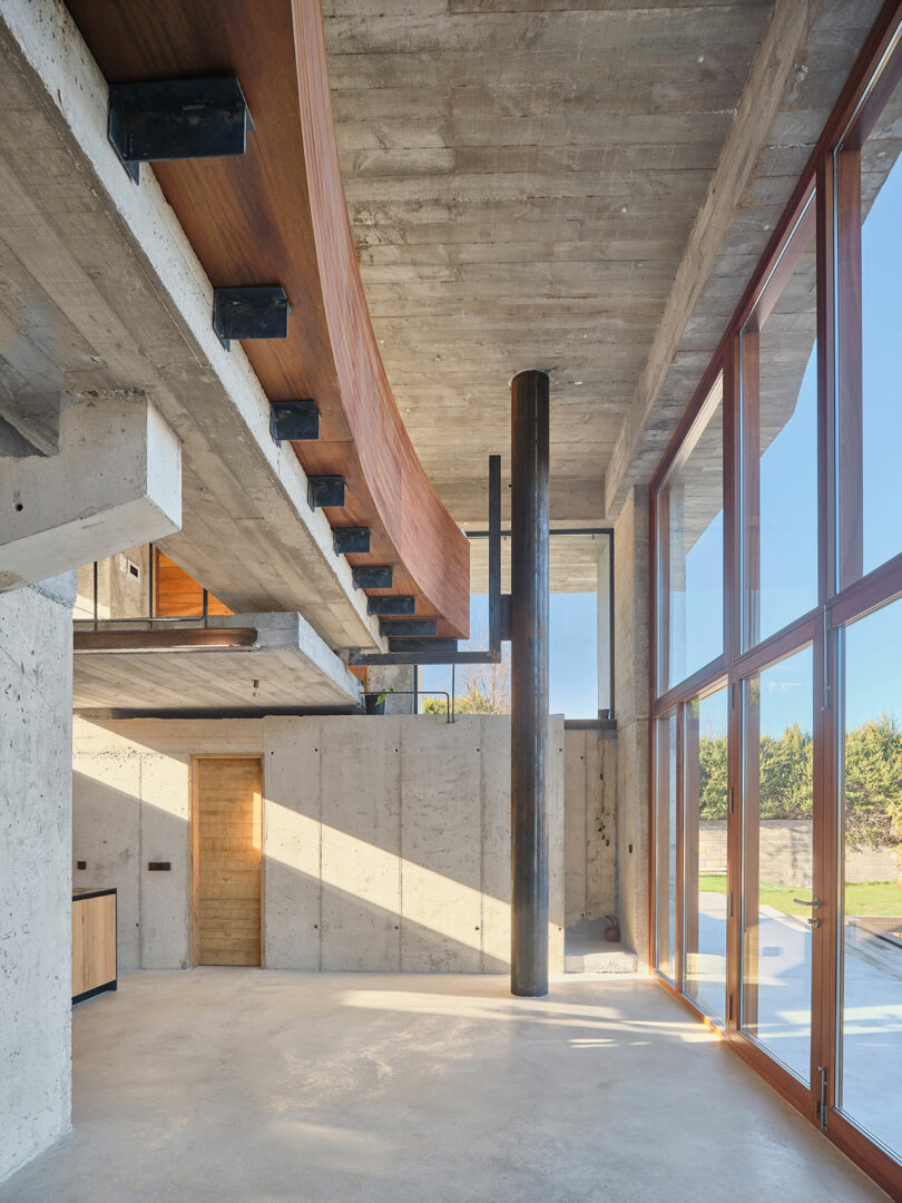 Modern interior with concrete walls, large windows, and a curved wooden element. Multilevel open space with visible beams, natural light streaming in, and outdoor view.