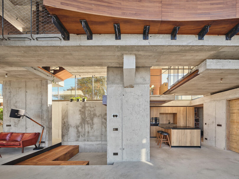 Modern open-plan interior with exposed concrete walls, wooden accents, a red sofa, and a black floor lamp. A kitchen area with stools is visible in the background. Large windows allow natural light.