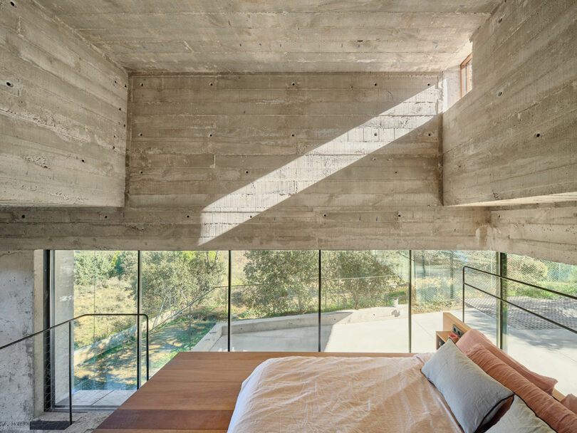 Interior of a modern room with concrete walls, large windows, a wooden platform bed, and nature views outside.