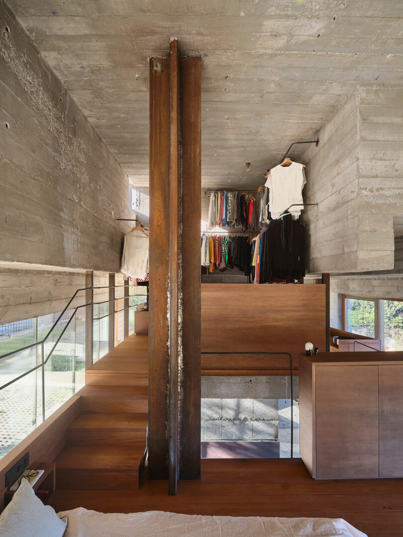 Modern interior with concrete walls, wooden flooring, and a central steel beam. An open wardrobe with hanging clothes is visible on the upper level, with stairs and railings on the left.