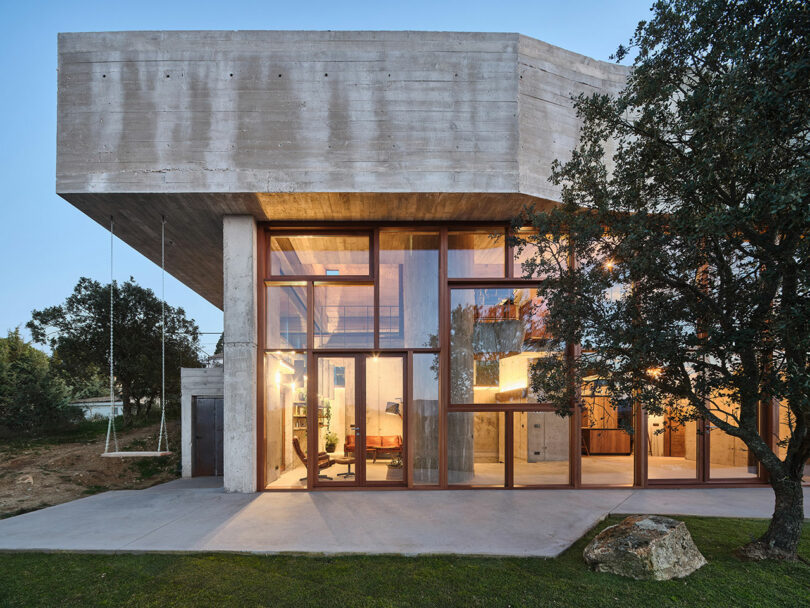 Modern two-story building with large glass windows and concrete structure, surrounded by trees and grass. A swing hangs from the structure.