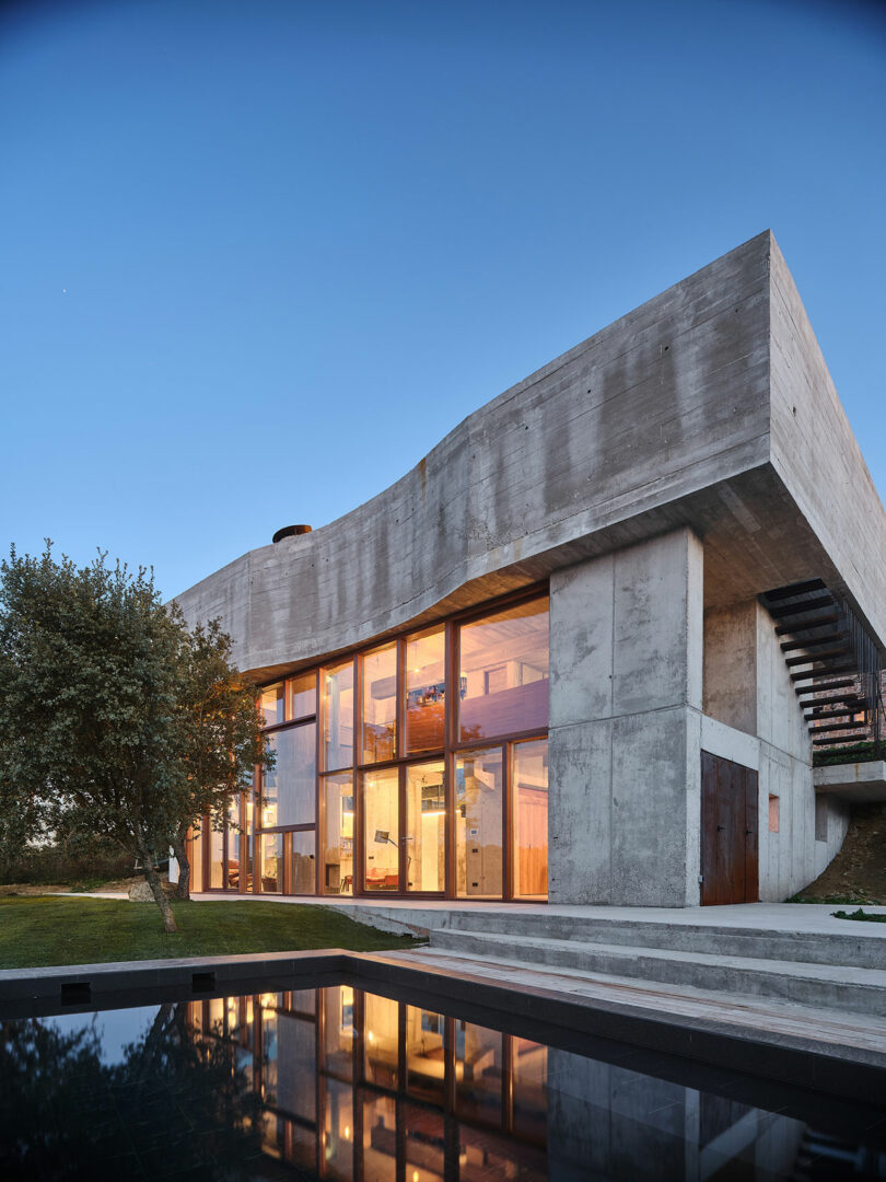 Modern concrete building with large glass windows and an exterior staircase, adjacent to a reflecting pool and a tree.