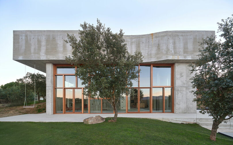 Modern concrete building with large wooden-framed windows, surrounded by grass and trees.