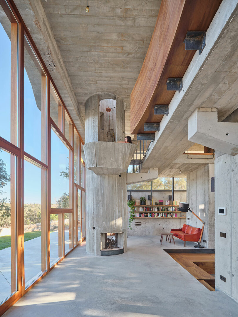 Spacious modern living area with large windows, concrete walls, a central fireplace, wooden and metal stairs, a bookshelf, and a red chair. Outdoor scenery visible through the windows.