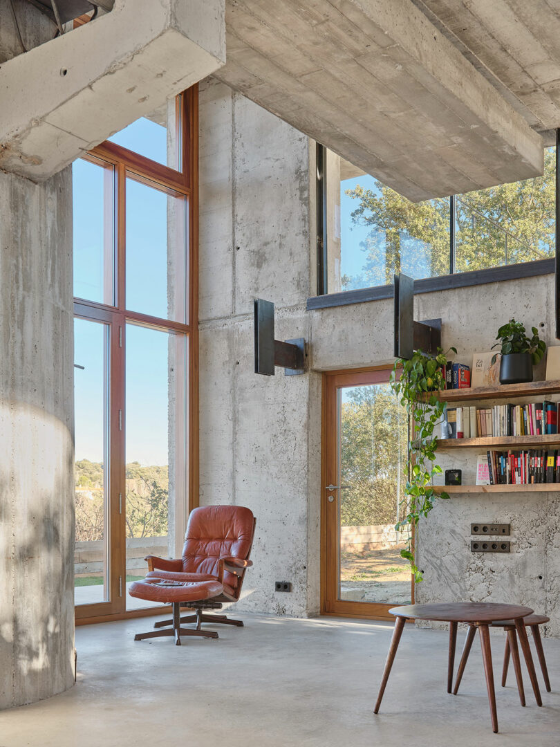 Modern industrial-style room with concrete walls, large windows, and wooden elements. Features a brown leather chair, bookshelf with plants, and a small table. Natural light illuminates the space.