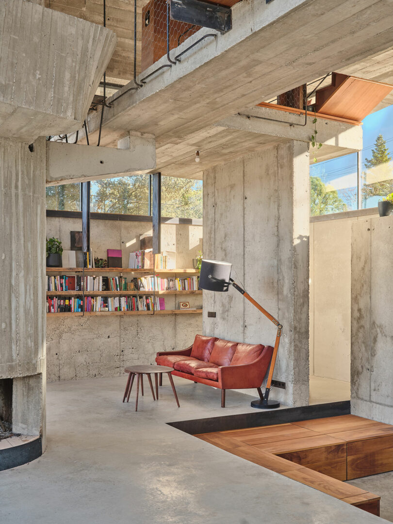 Industrial-style room with exposed concrete walls and ceiling. A leather sofa, floor lamp, and small table are on the concrete floor. Bookshelves line the walls with large, high windows above.